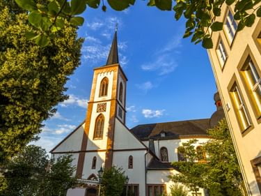 Weiße Kirche mit spitzem Turm und Uhr in Bitburg, eingerahmt von grünen Bäumen und angrenzenden gelben Gebäuden unter blauem Himmel.