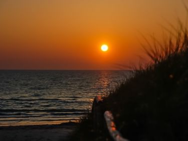 Orange Sonne über der Ostsee mit ruhigem Wasser und einer Düne mit Gras im Vordergrund.