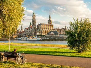 Fahrrad am Elberadweg mit Blick auf Dresdens historische Altstadt mit barocken Kirchen und Türmen, eingerahmt von herbstlichen Bäumen.