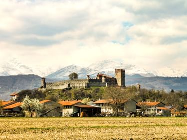 Mittelalterliche Burg Castello di Pavone auf einem Hügel mit Dorfhäusern darunter und schneebedeckten Alpen im Hintergrund unter bewölktem Himmel.