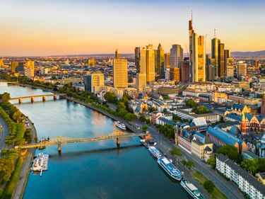Luftaufnahme der Frankfurter Skyline mit modernen Wolkenkratzern und historischen Gebäuden am Main bei Sonnenuntergang. Brücken und Schiffe sind sichtbar.