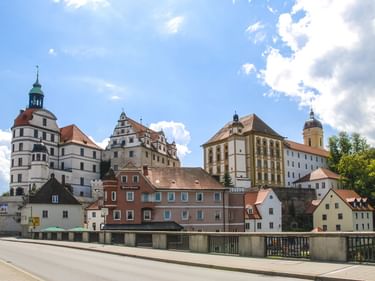 Historische Gebäude in Neuburg an der Donau mit Schlosstürmen, Kirchtürmen und bunten Fassaden entlang der Donau unter blauem Himmel.