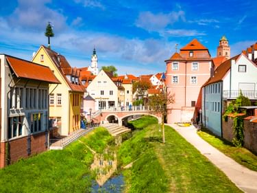Fischerplatz in Donauwörth mit bunten historischen Gebäuden, grünem Kanal und Kirchtürmen unter blauem Himmel.