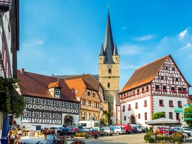 Bayerischer Marktplatz mit bunten Fachwerkhäusern und Kirche mit hohem Turm. Autos parken auf dem Platz unter blauem Himmel.