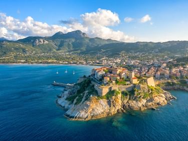 Luftaufnahme der historischen Festung Calvi auf einer Felshalbinsel auf Korsika, mit blauem Mittelmeer, Segelbooten und Bergen im Hintergrund.