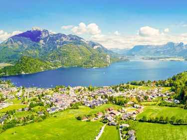 Luftaufnahme von St. Gilgen mit grünen Wiesen am Wolfgangsee, umgeben von Bergen unter blauem Himmel mit weißen Wolken.