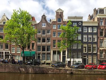 Reihe traditioneller Amsterdamer Grachtenhäuser mit verschiedenen Giebelfassaden entlang eines Kanals. Bäume säumen das Ufer, wo mehrere Autos parken.