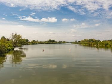 Drei Stand-up-Paddler auf dem ruhigen Altmühlsee bei Gunzenhausen. Grüne Bäume säumen die Ufer unter blauem Himmel mit weißen Wolken.