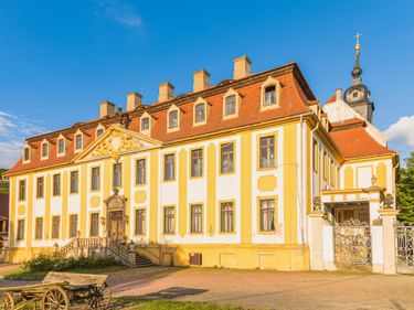 Barockschloss in Diesbar-Seußlitz mit gelb-weißer Fassade, rotem Mansarddach und Kirchturm. Ein alter Holzwagen steht im Innenhof.