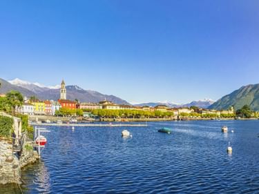Blick auf die Uferpromenade von Ascona am Lago Maggiore mit bunten Gebäuden, Kirchturm, Booten auf blauem Wasser und schneebedeckten Bergen.