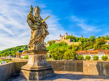 Bronzestatue des Heiligen Kilian auf steinerner Brücke in Würzburg mit Festung Marienberg auf dem Hügel und Main im Hintergrund unter blauem Himmel.