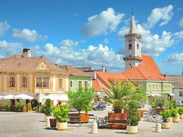 Bunter Stadtplatz in Rust mit historischen Gebäuden, weißem Kirchturm und Pflanzkübeln mit Bänken unter blauem Himmel mit weißen Wolken.