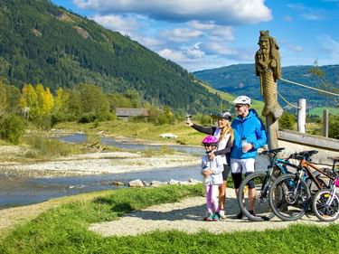 Familie mit drei Fahrrädern posiert neben einer Holzskulptur an der Mur, mit grünen Bergen und blauem Himmel im Hintergrund.