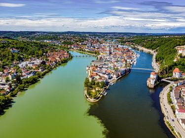 Luftaufnahme von Passau mit der Altstadthalbinsel zwischen zwei Flüssen mit Brücken, bunten Gebäuden und grünen Wäldern um die Stadt.