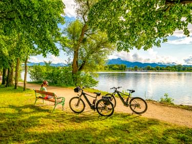 Person entspannt auf einer Bank am Chiemsee mit zwei abgestellten Fahrrädern. Grüne Bäume rahmen den Blick auf den See und die Berge in Bayern.