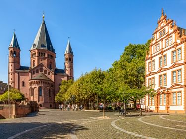 Mainzer Dom mit roten Sandsteintürmen neben einem Kopfsteinpflasterplatz. Ein orangefarbenes Barockgebäude steht rechts, grüne Bäume säumen den Platz.