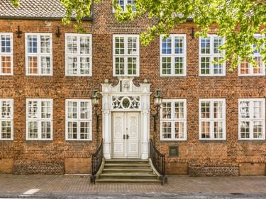 Drohses Hus: symmetrisches rotes Backsteingebäude mit weißen Fenstern, verziertem weißen Eingangsportal mit Treppe und grünem Laub.
