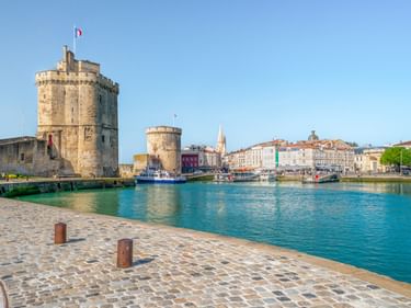 Alter Hafen von La Rochelle mit mittelalterlichen Steintürmen am türkisfarbenen Hafenwasser. Kopfsteinpflaster im Vordergrund, historische Gebäude am Ufer.