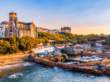 Küstenansicht von Biarritz mit gotischer Kirche auf einem Hügel, weißen Gebäuden, kleinem Hafen mit Booten und felsiger Küste bei Sonnenuntergang.