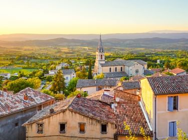 Luftaufnahme von Bonnieux mit Terrakottadächern, Kirchturm und hügeliger Provence-Landschaft bei Sonnenuntergang in goldenem Licht.