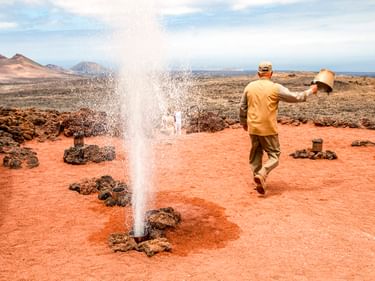 Parkranger demonstriert geothermische Aktivität im Timanfaya-Nationalpark, Lanzarote. Wasser spritzt als Geysir aus rotem Vulkanboden.