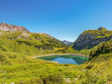 Formarinsee, ein türkisfarbener Bergsee umgeben von grünen Almwiesen und felsigen Gipfeln unter blauem Himmel in den Alpen.
