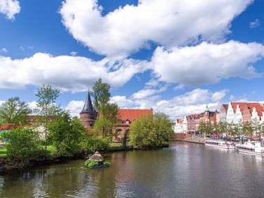Flussansicht von Lübeck mit mittelalterlichem Turm, bunten Giebelhäusern entlang des Ufers und grünen Bäumen unter blauem Himmel mit weißen Wolken.