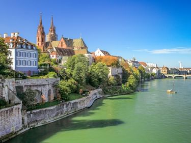 Basler Altstadt am Rhein mit den Doppeltürmen des Basler Münsters, historischen Gebäuden und einer Brücke unter blauem Himmel.