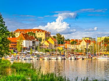 Hafen in Ryn mit Segelbooten am Ufer. Bunte Gebäude mit roten Dächern säumen den Hafen unter blauem Himmel mit weißen Wolken.