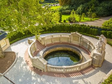 Luftaufnahme des kreisförmigen Donauquelle-Denkmals in Donaueschingen mit verzierter Steinbalustrade, Skulpturen und Wasserbecken umgeben von Park.
