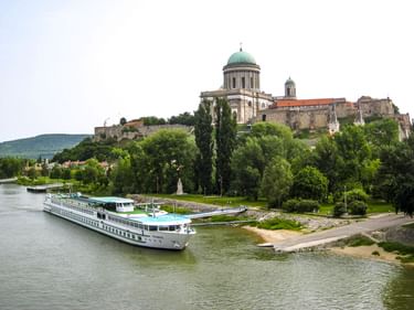 Weißes Flusskreuzfahrtschiff auf der Donau mit Basilika mit grüner Kuppel und historischen Gebäuden auf einem Hügel, umgeben von Bäumen.