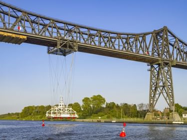 Rendsburger Schwebefähre mit Stahlfachwerk über einem Kanal. Eine hängende Fährplattform befindet sich unter der Brücke. Rote Bojen markieren die Wasserstraße.