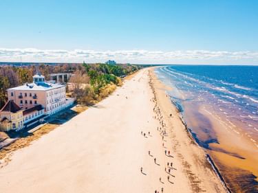 Luftaufnahme des breiten Sandstrands von Jurmala an der Ostsee. Menschen spazieren am Ufer, historische Gebäude und Wald links sichtbar.