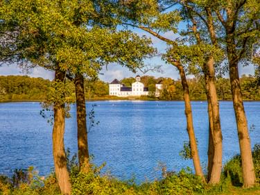 Weißes Schloss Gravenstein über einem blauen See, eingerahmt von Bäumen mit grünem Laub in warmem Sonnenlicht unter klarem Himmel.