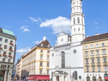 Horse-drawn carriages on cobblestone square in front of white church with tall spire. Historic buildings surround the plaza under blue sky.