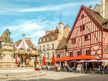 Place François Rude in Dijon mit historischem Brunnen, Karussell und roten Fachwerkhäusern. Café-Terrassen mit roten Sonnenschirmen.