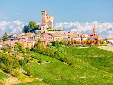 Hügelstadt Alba in Italien mit mittelalterlichen Türmen und Kirchturm, umgeben von grünen Weinbergen unter blauem Himmel mit weißen Wolken.