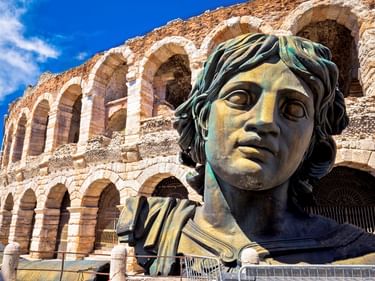 Große Bronzekopfskulptur mit welligem Haar vor dem antiken römischen Amphitheater Arena di Verona mit seinen charakteristischen Steinbögen.
