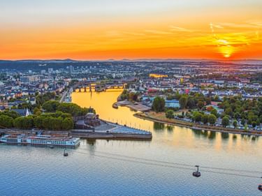 Aerial view of Deutsches Eck in Koblenz at sunset, showing the Rhine and Moselle rivers converging. Cable cars cross the river, with cityscape and mountains.
