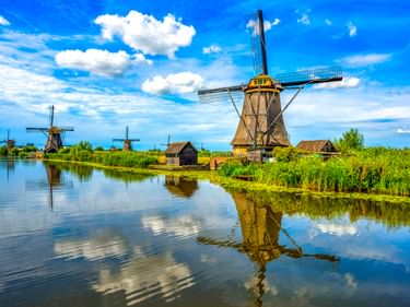 Traditionelle niederländische Windmühlen in Kinderdijk spiegeln sich in einem ruhigen Kanal unter blauem Himmel mit weißen Wolken und grünem Schilf.