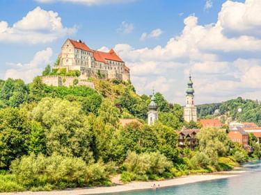 Burg Burghausen mit roten Dächern auf einem Hügel über einer Stadt am Fluss mit Kirchtürmen, umgeben von grünen Bäumen unter blauem Himmel.