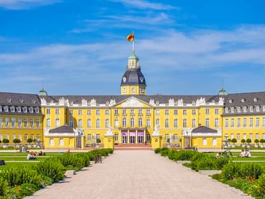 Gelbes barockes Karlsruher Schloss mit Mittelturm und Flagge, vom formalen Garten mit geschnittenen Hecken und Kiesweg unter blauem Himmel.