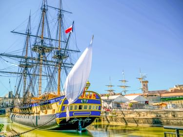 Historische Fregatte Hermione mit blau-gelbem Rumpf, drei Masten und französischer Flagge im Hafenbecken von Rochefort unter blauem Himmel.