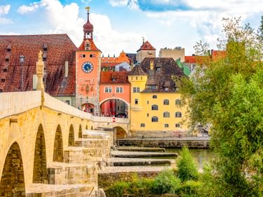 Stone Bridge in Regensburg with medieval arches spanning the Danube River. Colorful historic buildings and a clock tower visible in the old town.