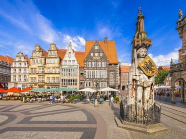 Bremer Marktplatz mit der Roland-Statue im Vordergrund und bunten historischen Giebelhäusern hinter Straßencafés unter blauem Himmel.