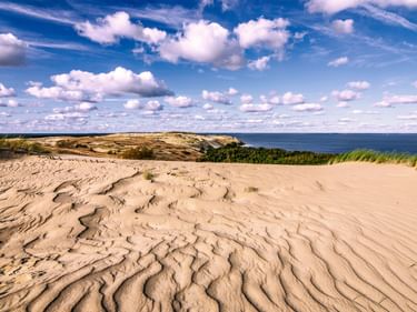 Parnidis-Düne in Litauen mit gewellten Sandmustern im Vordergrund, grüner Vegetation und blauer Ostsee unter bewölktem Himmel.