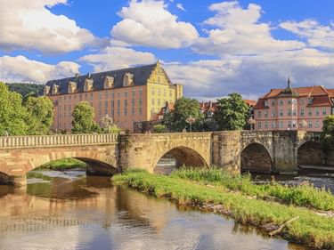Stone bridge crossing river with Welfenschloss palace and historic buildings in Hann. Münden under blue sky with white clouds.