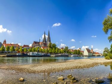 Panoramablick auf die Regensburger Altstadt vom Donauufer mit Domtürmen, historischen Gebäuden und Schiffen auf dem Wasser unter blauem Himmel.