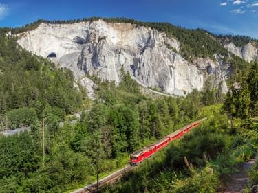 Roter Zug fährt durch die Rheinschlucht Ruinaulta mit weißen Kalksteinfelsen und dichtem Wald. Klarer blauer Himmel über den Felsformationen.