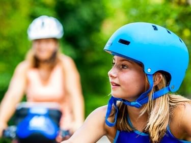 Young girl wearing a blue cycling helmet looks ahead with an adult cyclist blurred in the background among green foliage.